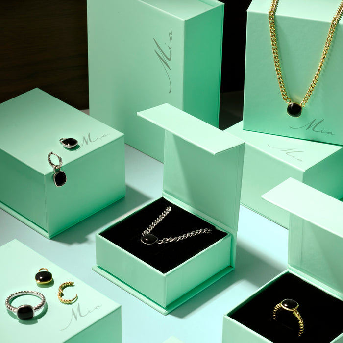 Black gemstone jewelry collection in stainless steel featuring pendant necklaces, rings and earrings displayed in mint green Mia jewelry boxes, elegant modern jewelry with polished black stones.