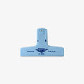 A light blue plastic binding clip with the brand name 'Pencas' and model 'SLAMPS' printed in dark blue on the front, set against a plain light gray background.