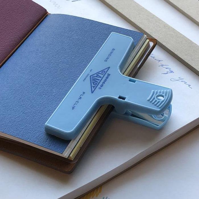 A light blue plastic binder clip holding a blue notebook on top of a white page with handwritten text, photographed in a styled flat-lay arrangement.