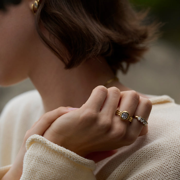 Close-up lifestyle shot of a woman's hands wearing a gold ring with a round clear stone and ridged band, paired with a stone-set gold ring and a cream knit sweater.

Photo lifestyle en gros plan des mains d'une femme portant une bague dorée sertie d'une pierre transparente ronde à anneau strié, agencée avec une bague dorée sertie de pierres et un tricot crème.