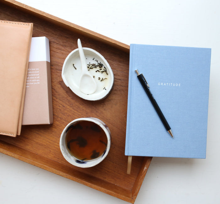 A light blue hardcover notebook with the word 'GRATITUDE' printed on the cover in capital letters. table setup with tea and pen.