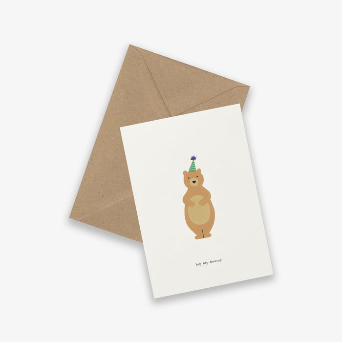 A birthday greeting card with an illustration of a bear wearing a party hat, with the text 'Hip Hip Hooray'. It is presented with a brown kraft envelope.