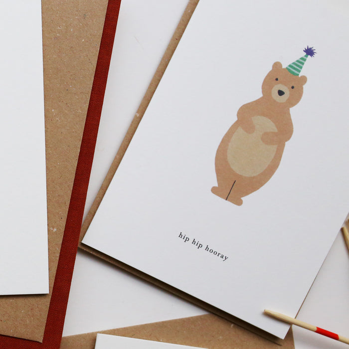 A birthday greeting card with an illustration of a bear wearing a party hat, with the text 'Hip Hip Hooray'. It is presented with a brown kraft envelope.