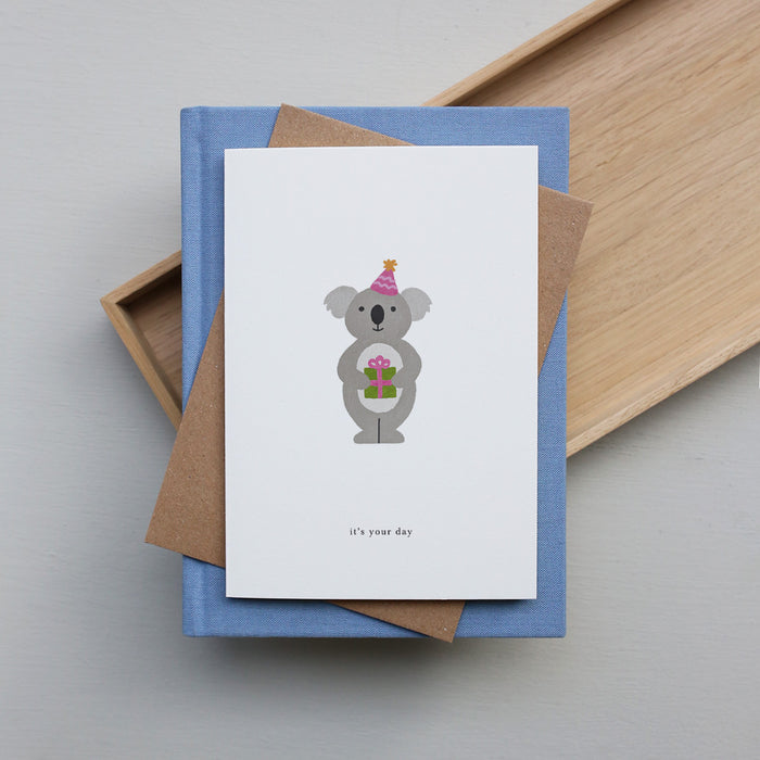 Greeting card with a koala illustration and 'it's your day' text, placed on a stack of books.