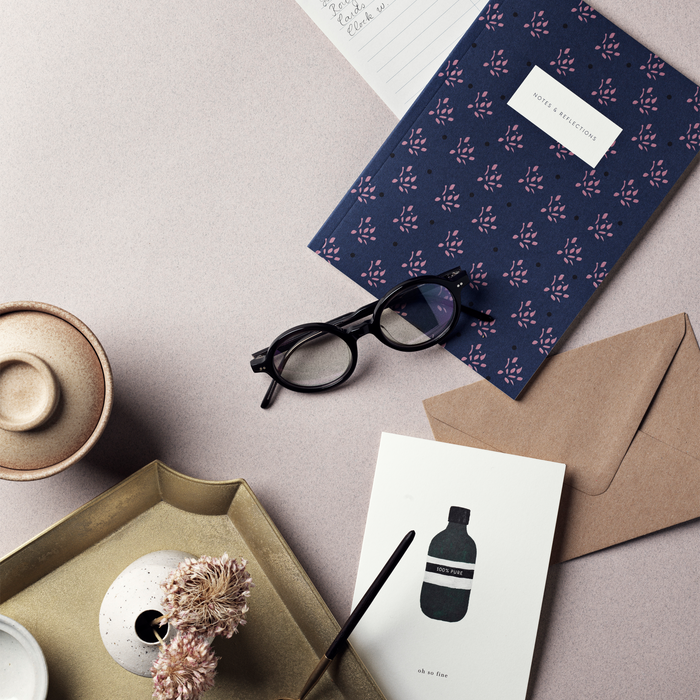 Table set up with stationary products including a simple illustrated greeting card with a bottle design with the text 'oh so fine' accompanied by a brown kraft envelope, displayed on a white background.