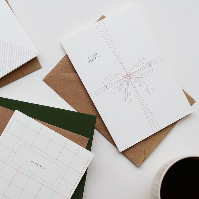 A simple white A6 greeting card with the text 'SIMPLY PERFECT' and a graphic of a bow, accompanied by a brown kraft envelope.