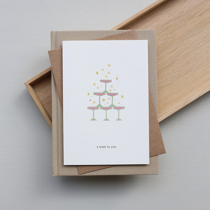 Greeting card with a cake illustration and 'A toast to you' text, placed on a brown envelope with a wooden tray in the background.