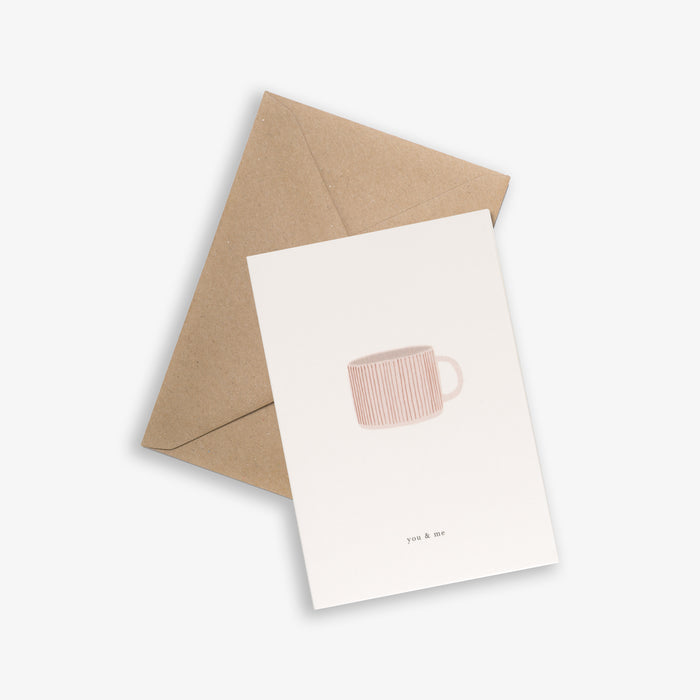 A greeting card with a illustration of a cup on it and a text 'you & me', accompanied by a brown kraft envelope, placed on a white background.