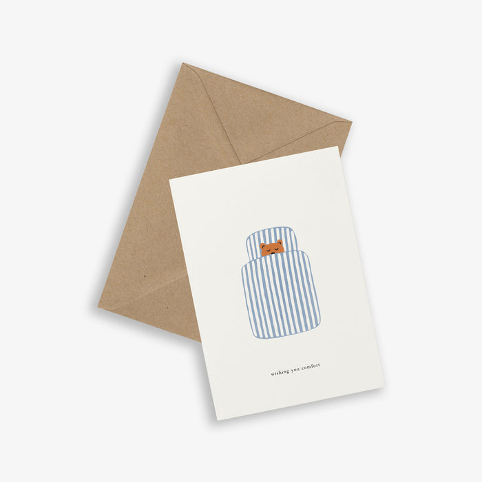 A greeting card with a illustration of a blue and white striped duvet with a sleeping bear and the text 'wishing you comfort', placed on a white background.