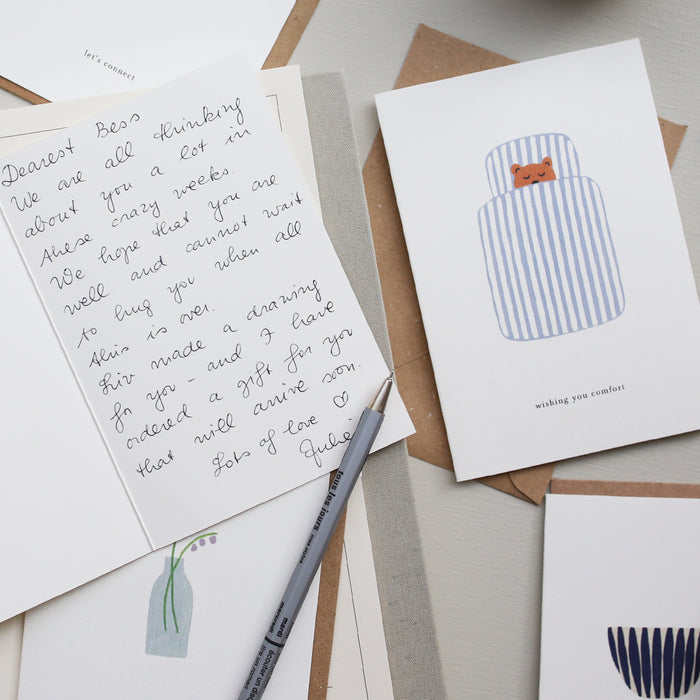 A greeting card with an illustration of a blue and white striped duvet with a sleeping bear and the text 'wishing you comfort'. next to a written card.