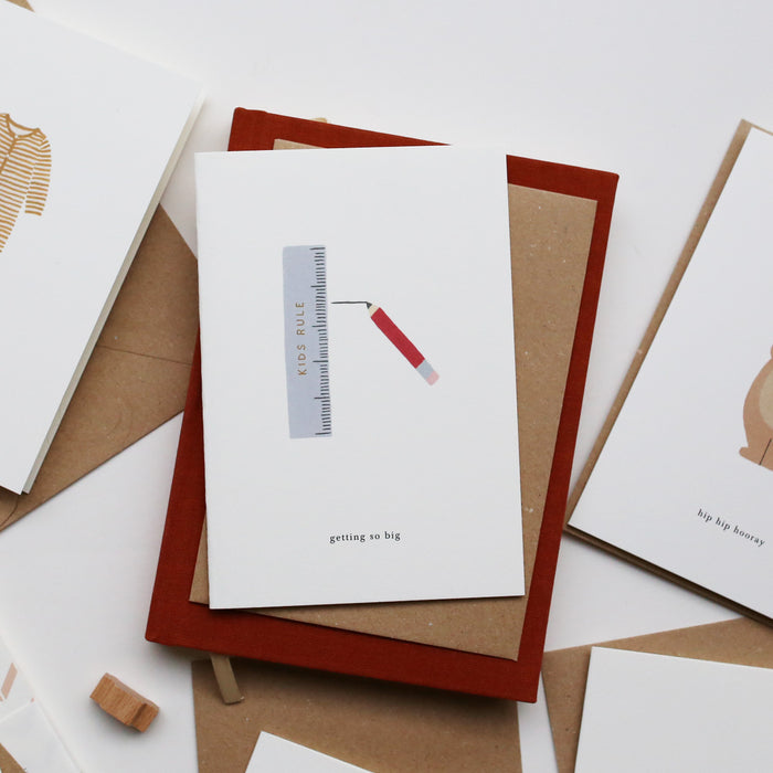 A greeting card with an illustration of a ruler and a pencil and with the text 'getting so big', accompanied by a brown kraft envelope, on a white background.