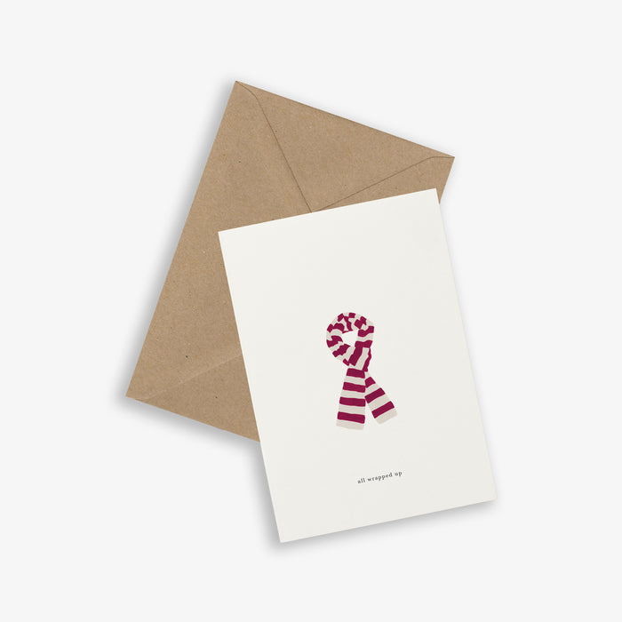 A simple illustrated greeting card with a scarf design with the text 'all wrapped up' accompanied by a brown kraft envelope, displayed on a white background.