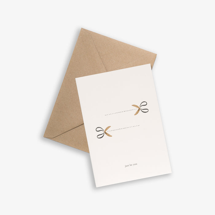 A simple illustrated greeting card with two scissors and the text 'just be you', accompanied by a brown kraft envelope.