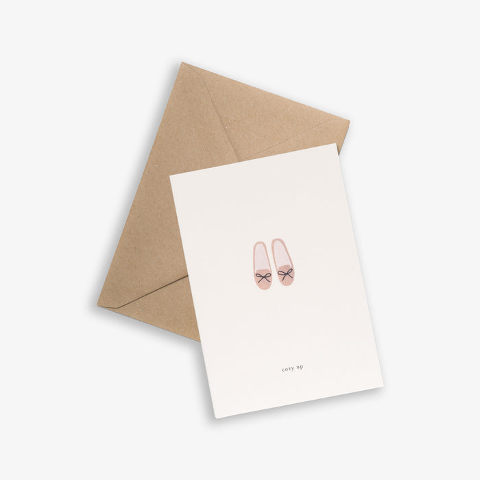 A greeting card with a simple illustration of slippers at the bottom, with a brown kraft envelope placed behind it. The card has a minimalist white background and text that reads 'cozy up'.