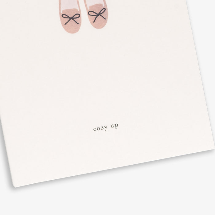 A greeting card with a simple illustration of slippers at the bottom. The card has a minimalist white background and text that reads 'cozy up'.