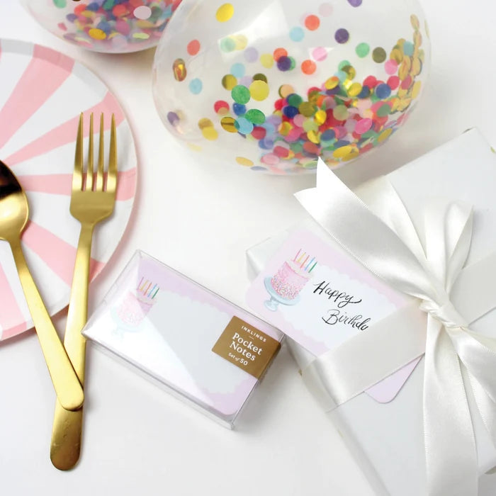 Table setting with birthday-themed items including a box of pocket notes, a fork, knife, and colorful confetti plates.