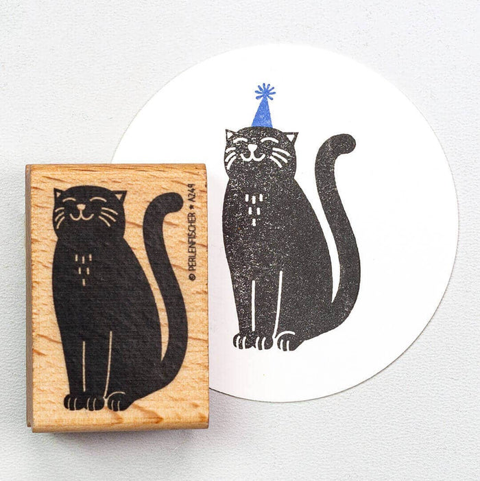 Rubber stamp with a black cat design on a wooden block, next to a printed card with a cat wearing a party hat.