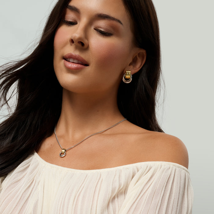A close-up of a woman wearing a delicate two-tone necklace and matching earrings, paired with an off-the-shoulder cream top against a light gray background.

Gros plan d'une femme portant un collier délicat deux tons et des boucles d'oreilles assorties, agencés avec un haut crème à épaules dénudées sur fond gris pâle.