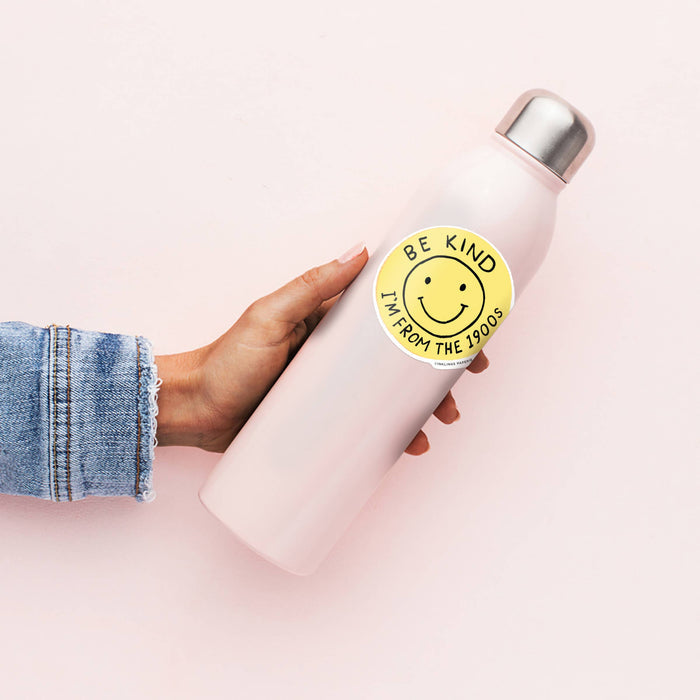 White water bottle with a yellow smiley face sticker and text, held by a person on a light pink background