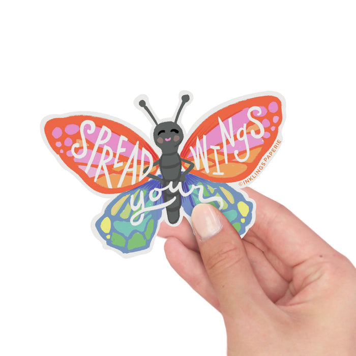 Butterfly-shaped sticker with 'Spread Wings' text held by a hand on a white background