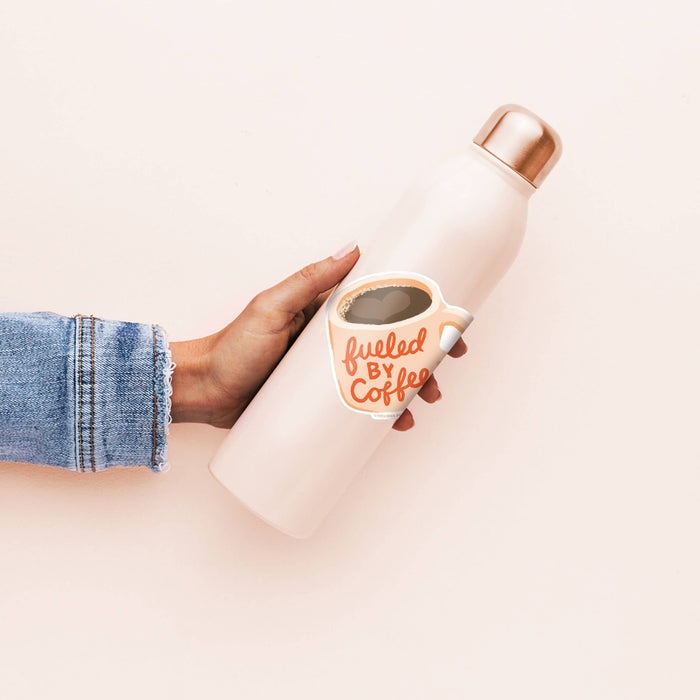 Person holding a white insulated bottle with a small cup labeled 'fueled by coffee' on a light pink background