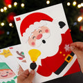 Hand holding a sheet of festive stickers with Santa Claus, reindeer, and snowflakes for windows against a blurred Christmas tree background.